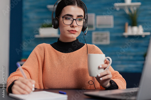 Student in online class holding cup looking at laptop screen and taking notes. Woman with headphones writing on notebook listening to university lecture. College girl with glasses studying remote. photo