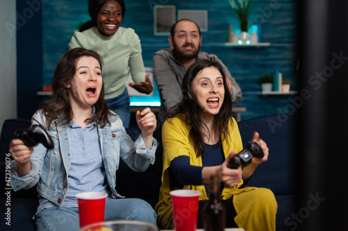 Happy smiling women playing videogames using gaming joystick winning online competition enjoying spending time in living room. Multi-ethnic friends having fun together during wekeend party