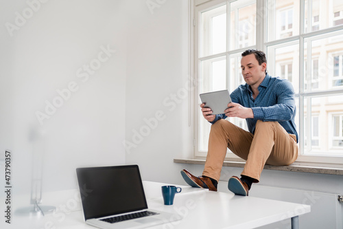 Male professional using digital tablet while sitting on window sill at office