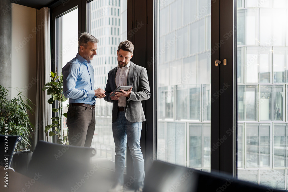 Businessmen discussing plan over digital tablet by window at work place Businessmen discussing plan over digital tablet by window at work place