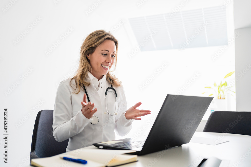 Smiling female doctor gesturing while talking during video call through laptop at desk Smiling female doctor gesturing while talking during video call through laptop at desk