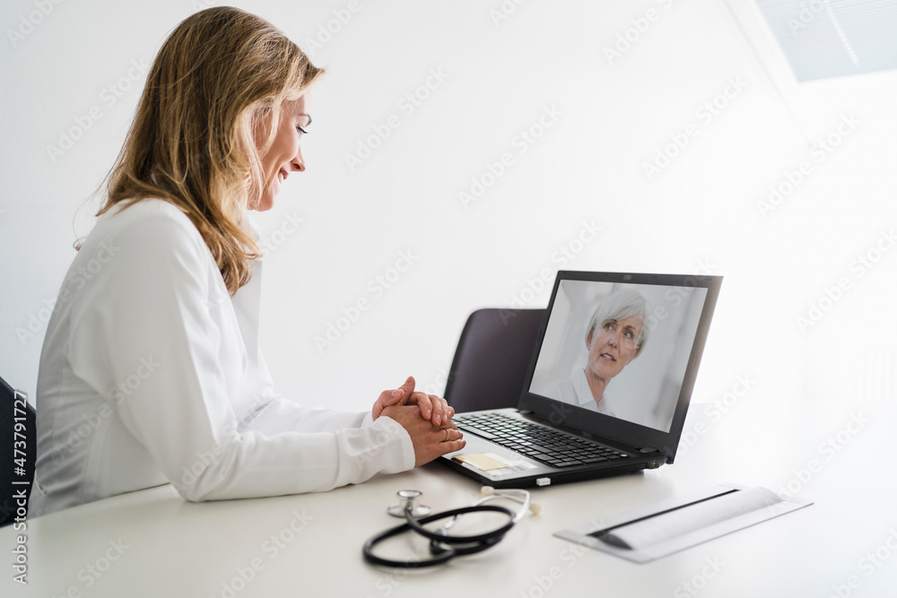 Smiling female doctor talking with senior patient during telemedicine through laptop at desk Smiling female doctor talking with senior patient during telemedicine through laptop at desk