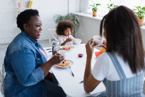 toddler african american girl having breakfast with smiling granny and blurred mom in kitchen