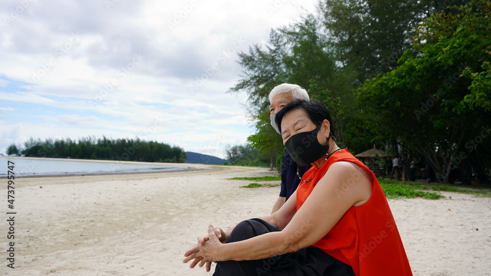 Asian senior couple with mask relax sitting on beach log white sand landscape and beautiful sea