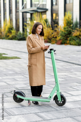 Happy woman in autumn coat using smartphone for renting modern electric scooter with an application, reads qr code with a mobile phone camera. Alternative city transport