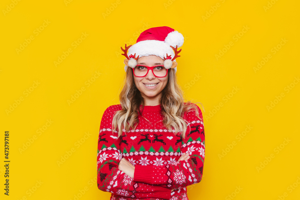 A young smiling blonde woman in glasses with deer antlers, a deer warm Christmas sweater and red Santa hat stands crossing her arms on her stomach isolated on color yellow background. New Year concept A young smiling blonde woman in glasses with deer antlers, a deer warm Christmas sweater and red Santa hat stands crossing her arms on her stomach isolated on color yellow background. New Year concept