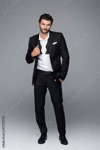 full length of bearded man in suit posing with hand in pocket on grey
