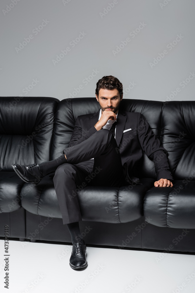 confident and bearded man in formal wear sitting on black couch on grey confident and bearded man in formal wear sitting on black couch on grey