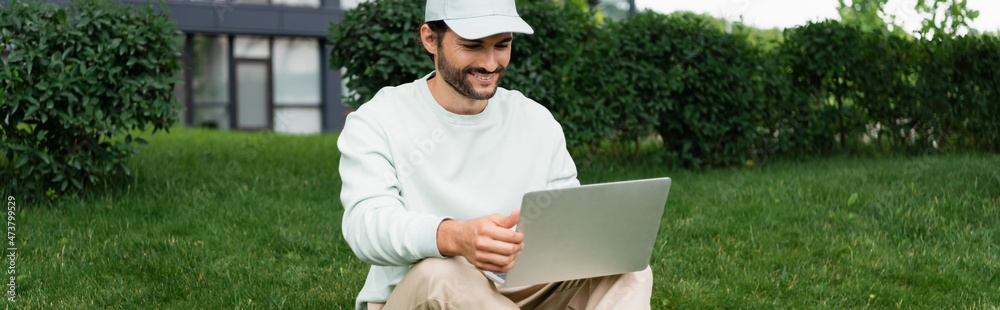 full length of happy freelancer using laptop while sitting on lawn, banner