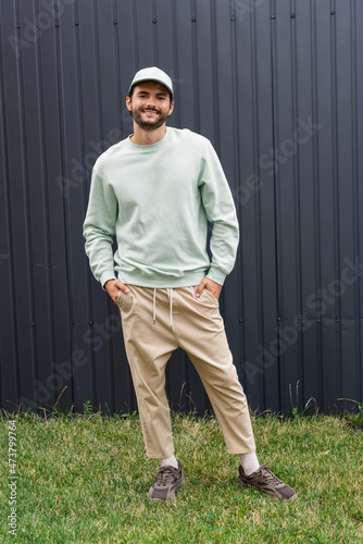 full length of smiling man in baseball cap posing with hands in pockets near metallic fence