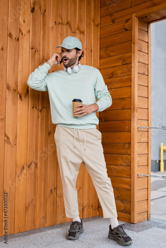 full length of shocked man in baseball cap holding coffee to go while talking on smartphone near building