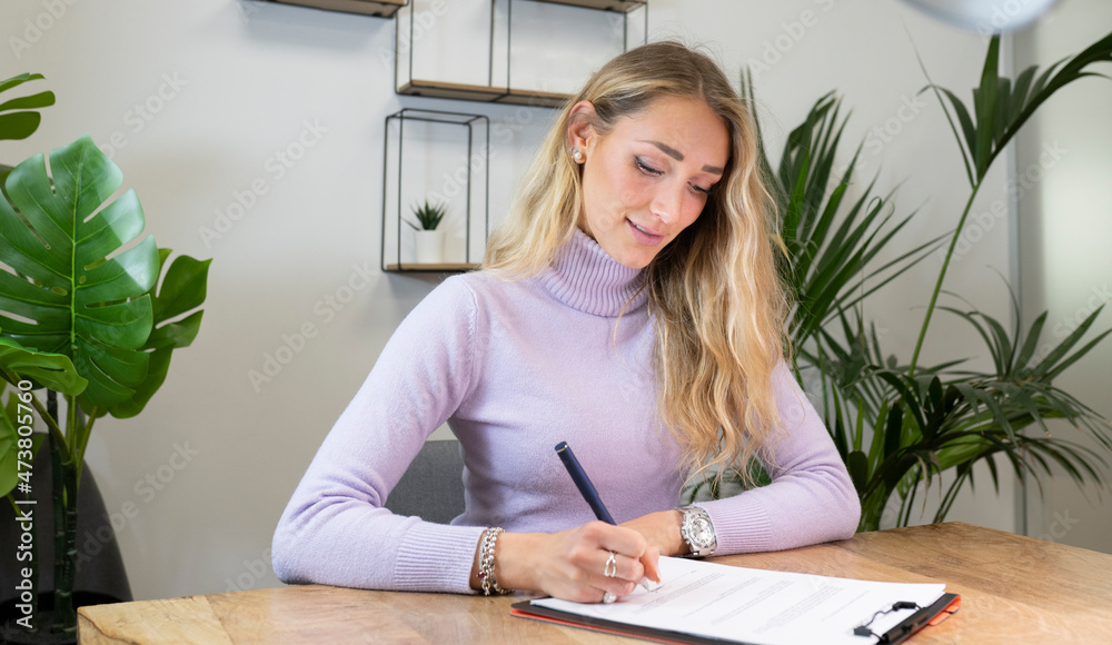 Blond businesswoman signing documents sitting at desk in office Blond businesswoman signing documents sitting at desk in office