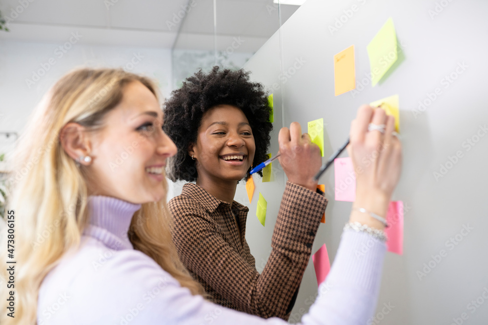 Smiling female colleagues writing on adhesive notes together while discussing in office
