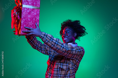 Portrait of happy young girl holding big red gift box isolated on dark green studio background in purple neon light.