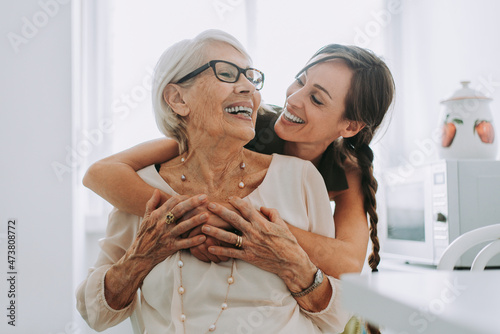 Cheerful woman hugging grandmother sitting in kitchen photo