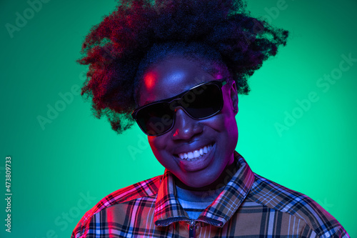 Close-up African young girl's portrait on dark green studio background in purple neon light. Afro hairdo. Concept of human emotions