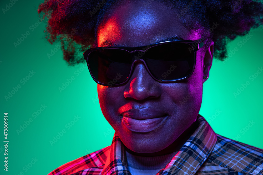 Close-up face of dark skinned young girl in sunglasses isolated on dark green studio background in purple neon light. Close-up face of dark skinned young girl in sunglasses isolated on dark green studio background in purple neon light.