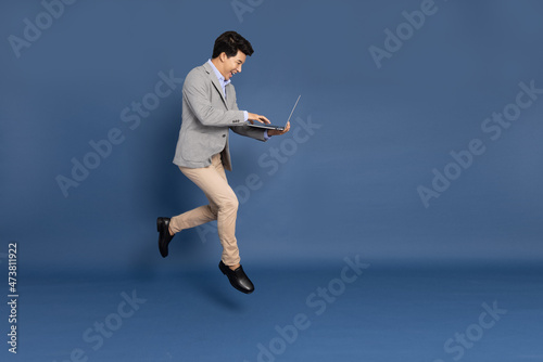 Fotografie, Obraz Young Asian business man jumping in air using laptop computer isolated on deep b
