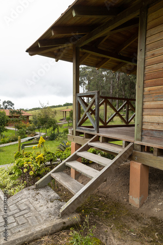 detail of the corner of a wooden cabin with balcony and steps, rustic house structure with gardin and trees in the background, decoration with nature, lifestyle and architecture