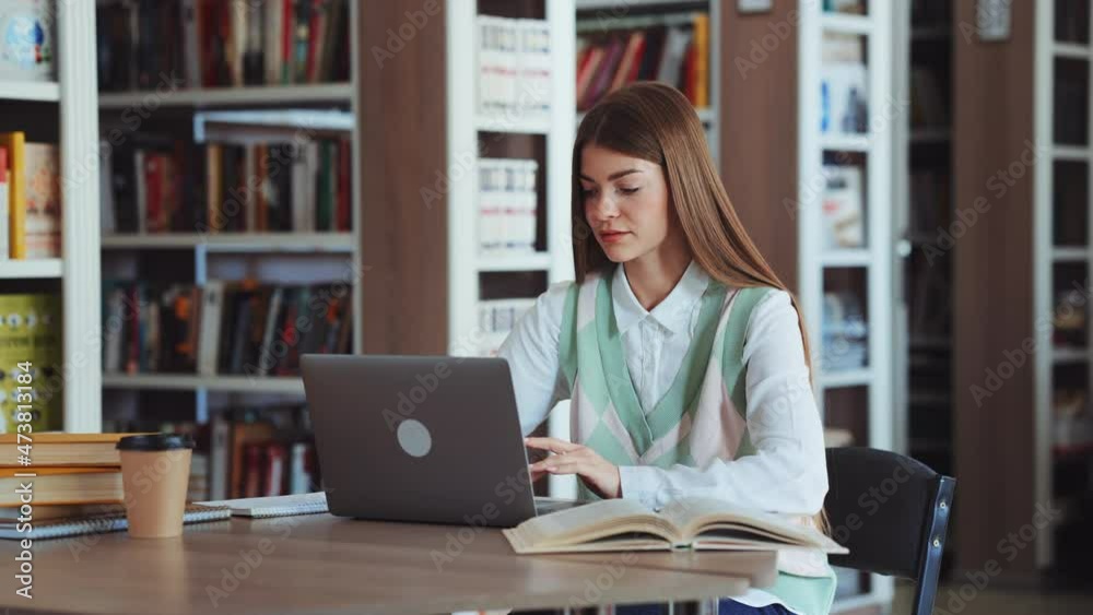 Female student with long hair working on laptop in library, making notes on paper, opened books lying on table, carton cup beside. Concept of education Female student with long hair working on laptop in library, making notes on paper, opened books lying on table, carton cup beside. Concept of education