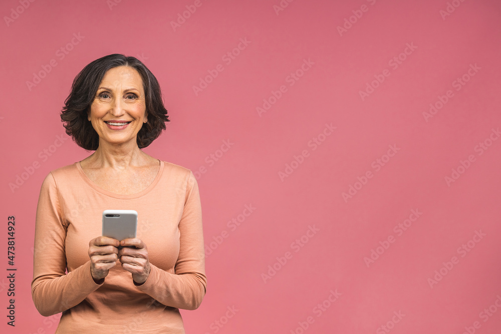 Happy mature senior woman holding smartphone using mobile online apps, smiling old middle aged lady texting sms message chatting on phone looking at cellphone isolated over pink background. Happy mature senior woman holding smartphone using mobile online apps, smiling old middle aged lady texting sms message chatting on phone looking at cellphone isolated over pink background.