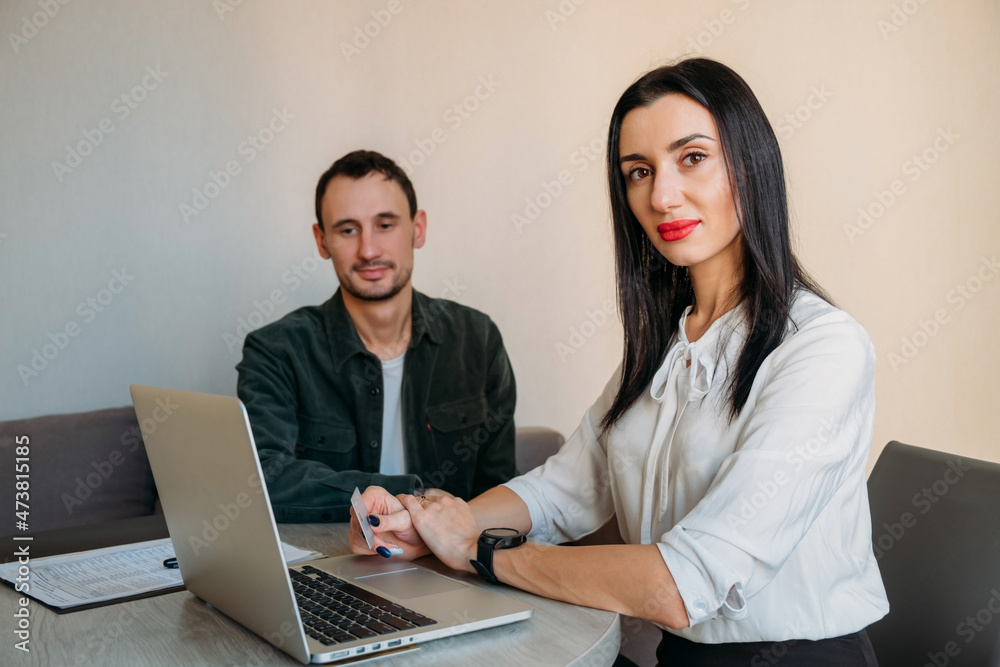 Business portrait of a beautiful female consultant sitting at the table and holding a client's credit card in her hand. In the background, a contented man sits on a couch. Business portrait of a beautiful female consultant sitting at the table and holding a client's credit card in her hand. In the background, a contented man sits on a couch.