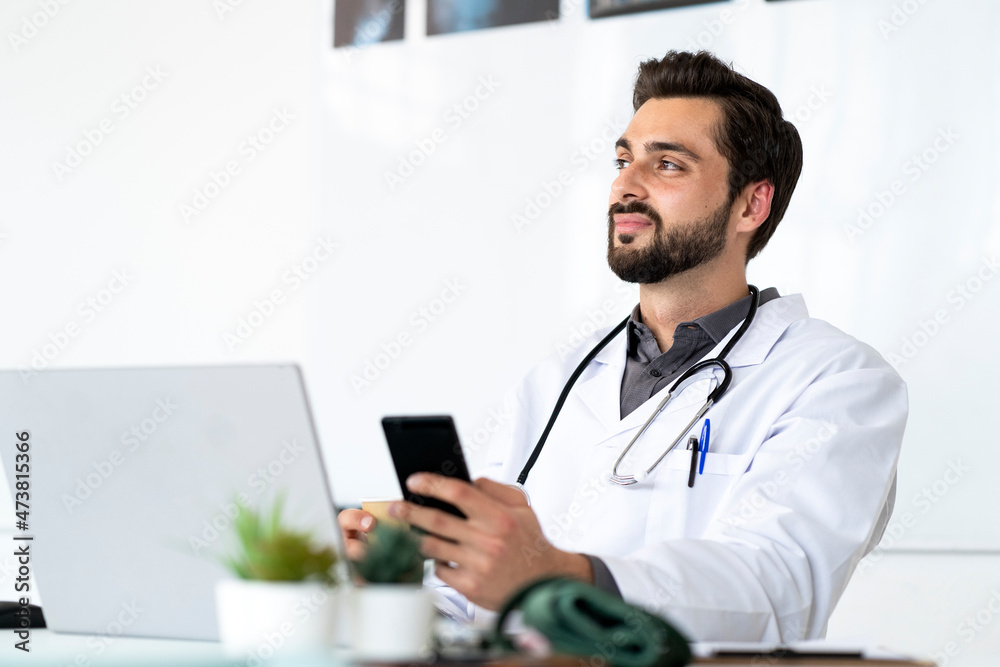 Male doctor with smart phone sitting at desk while looking away Male doctor with smart phone sitting at desk while looking away