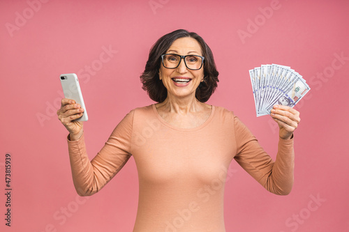 Happy winner! Image of mature aged happy senior woman standing isolated over pink background wall looking at camera holding money using mobile phone.