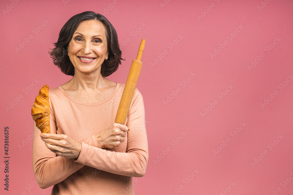 Senior mature aged woman baker holding a rolling pin and croissant, isolated on pink background. Senior mature aged woman baker holding a rolling pin and croissant, isolated on pink background.