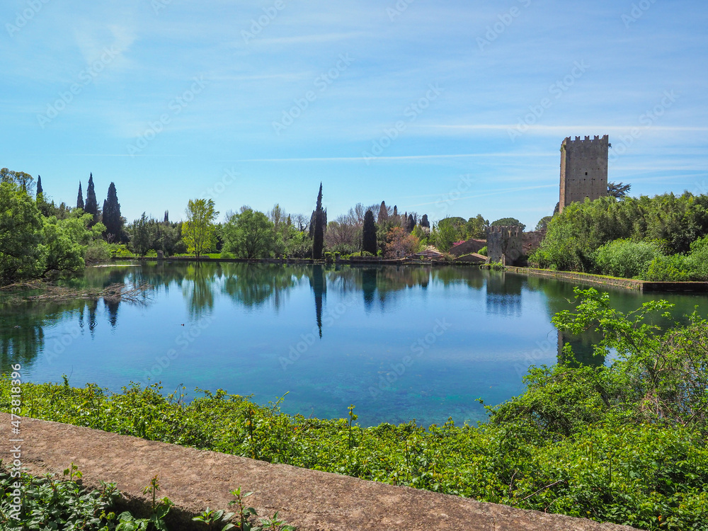 Fototapeta premium Garden of Ninfa or Giardino di Ninfa, with medieval castle ruins, near the lake and flowering plants of various colors. Amazing park of Caetani family during spring. Beautiful landscape with blue sky.