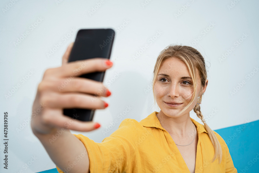 Smiling woman taking selfie through mobile phone by white wall Smiling woman taking selfie through mobile phone by white wall