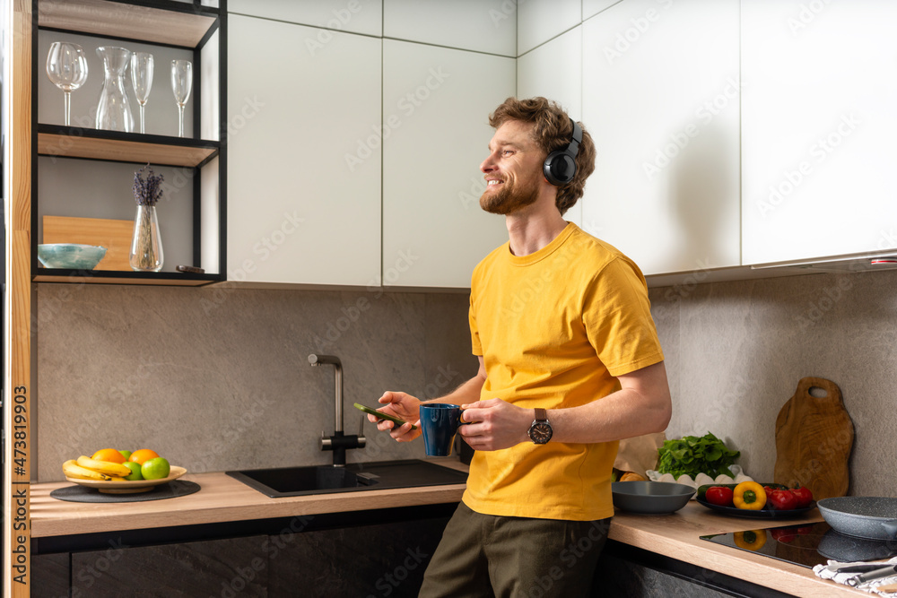 Smiling man holding coffee cup and smart phone while listening music through headphones in kitchen Smiling man holding coffee cup and smart phone while listening music through headphones in kitchen