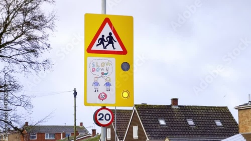 UK road sign indicating a school zone, possibility of children crossing the road, and a 20MPH speed limit. Flashing amber lights draw attention to the sign. The sign is located near a British school.