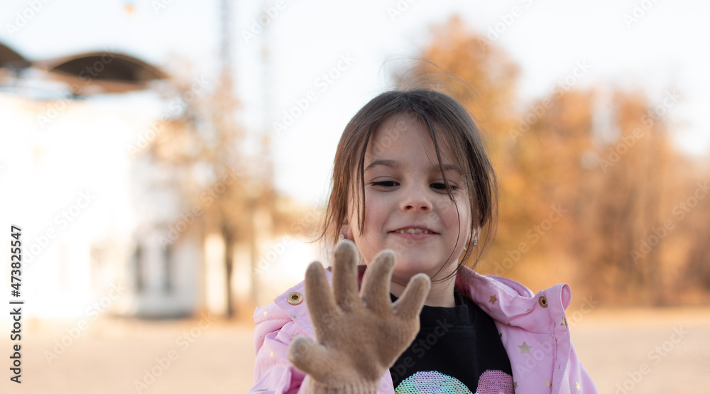 dark-haired little girl in a jacket with a hood smiling stretches out her hand in a glove standing on the autumn street dark-haired little girl in a jacket with a hood smiling stretches out her hand in a glove standing on the autumn street