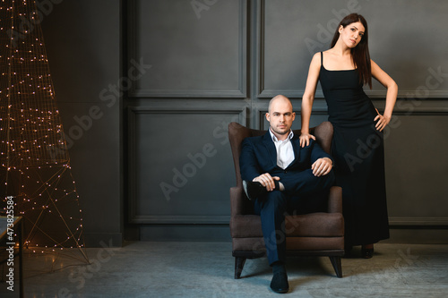 Man and woman in elegant outfits in a dark room with a Christmas tree. Girl with dark hair and a black dress with a bald guy in a suit sitting in a chair