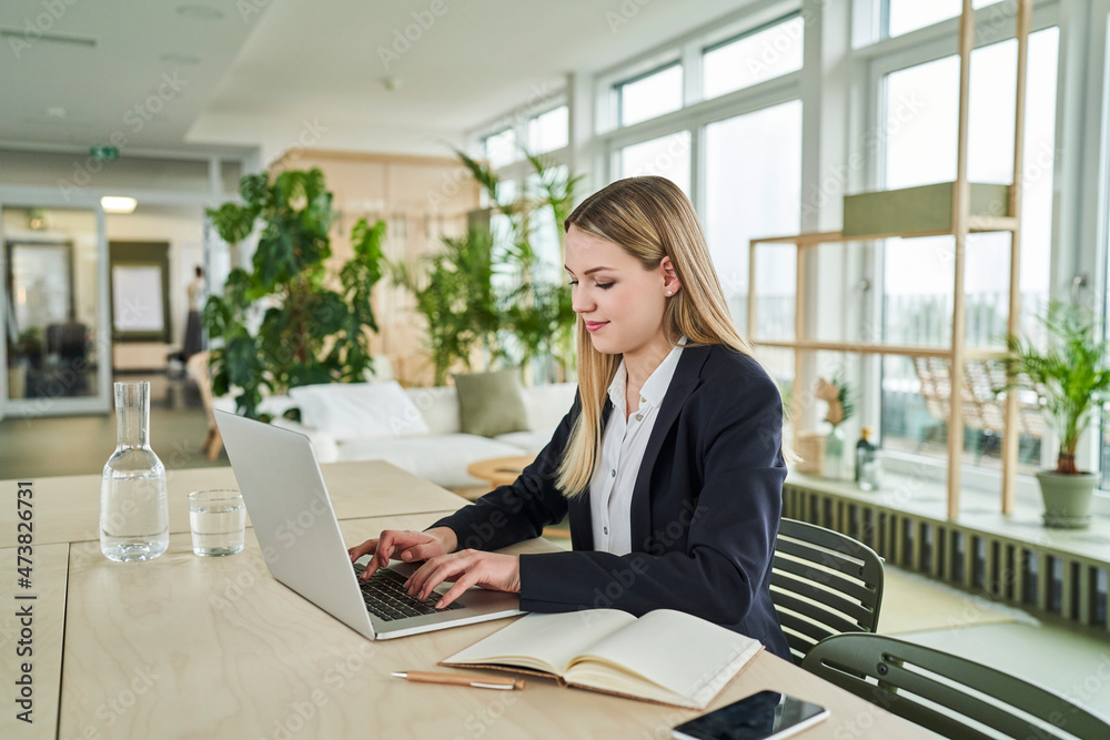 Blond trainee using laptop while sitting at desk in office Blond trainee using laptop while sitting at desk in office