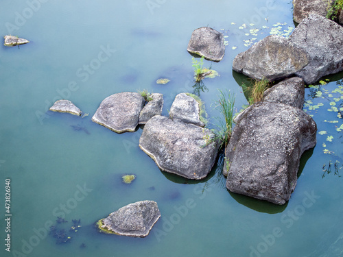 Top view of large stones in the river. Clean blue water is slightly turbid and has a milky hue. On the smooth whitish surface of the water there are few leaves of algae and duckweed.