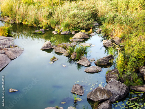 Amazing autumn nature: tall yellow dry grass and large stones near the riverbank. Plants are reflected on a calm surface of the water. Dim evening lighting.