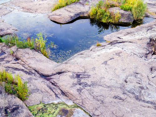 A water among flat huge stones. Gentle river bank. In some places tall grass and shrubs grow. Beautiful, picturesque autumn landscape. Part of the leaves dried and turned yellow.