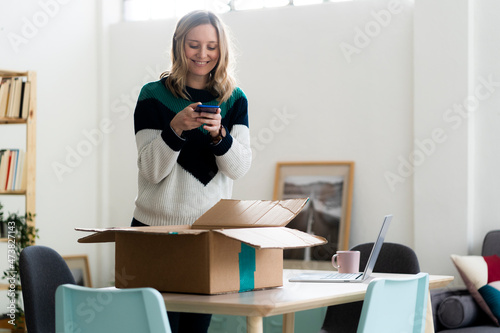 Smiling woman taking photo of package box through mobile phone at home photo