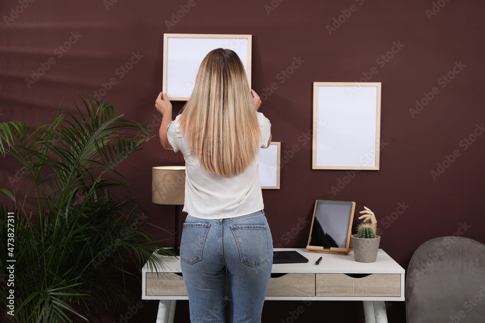 Woman hanging empty frame on brown wall indoors, back view. Mockup for design Woman hanging empty frame on brown wall indoors, back view. Mockup for design