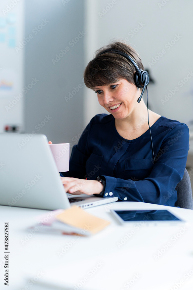 Smiling businesswoman wearing headset using laptop while working in office Smiling businesswoman wearing headset using laptop while working in office