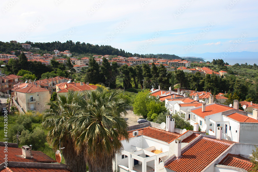 Top view of seaside town. Old traditional Greek houses with a red tiled roof, lots of trees and greenery, blue sea and mountains.