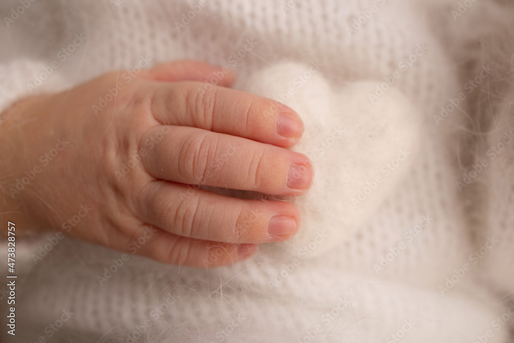 Tiny hand of a newborn baby. Soft hands of new born in white clothes hold white woolen heart. Fingers of a newborn baby hands close up. Studio macro photo. Happiness of a woman. Photography, concept. Tiny hand of a newborn baby. Soft hands of new born in white clothes hold white woolen heart. Fingers of a newborn baby hands close up. Studio macro photo. Happiness of a woman. Photography, concept.