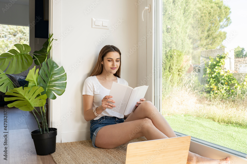 Young woman reading book near potted plant at home Young woman reading book near potted plant at home