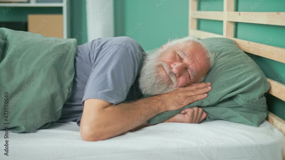 Portrait of an Elderly Man Sleeping Quietly on a Bed in His Room. Happy Retired Man Smiling, Lying in Bed, Sleeping on a Comfortable Mattress And Pillow, Resting Peacefully. Healthy Sleep Concept. Portrait of an Elderly Man Sleeping Quietly on a Bed in His Room. Happy Retired Man Smiling, Lying in Bed, Sleeping on a Comfortable Mattress And Pillow, Resting Peacefully. Healthy Sleep Concept.