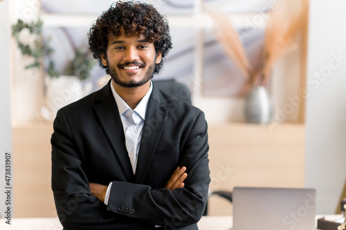 Business portrait of successful stylish young adult Indian businessman, manager or IT specialist, wearing a business suit, stands near his desk in the office with arms crossed, looks at camera, smiles