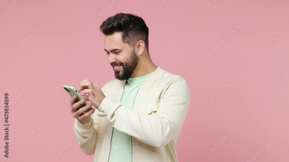 Young bearded brunet man 20s wears white shirt hold use mobile cell phone typing browsing swipe chatting send sms doing online shopping isolated on plain pastel light pink background studio portrait Young bearded brunet man 20s wears white shirt hold use mobile cell phone typing browsing swipe chatting send sms doing online shopping isolated on plain pastel light pink background studio portrait