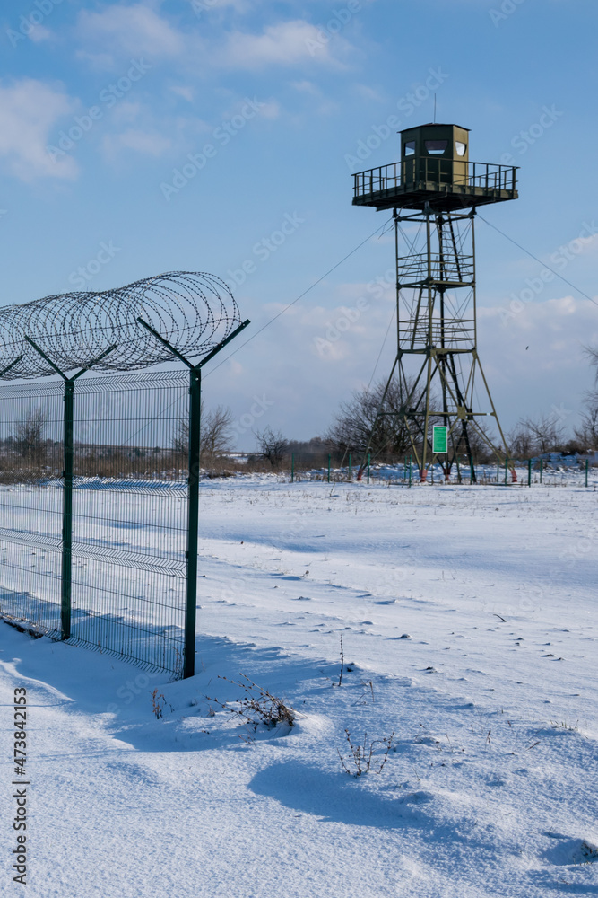Border fence with barbed wire in front of the surveillance tower in a snow-covered field against a blue sky. Border protection and counteraction to illegal migration concept Border fence with barbed wire in front of the surveillance tower in a snow-covered field against a blue sky. Border protection and counteraction to illegal migration concept