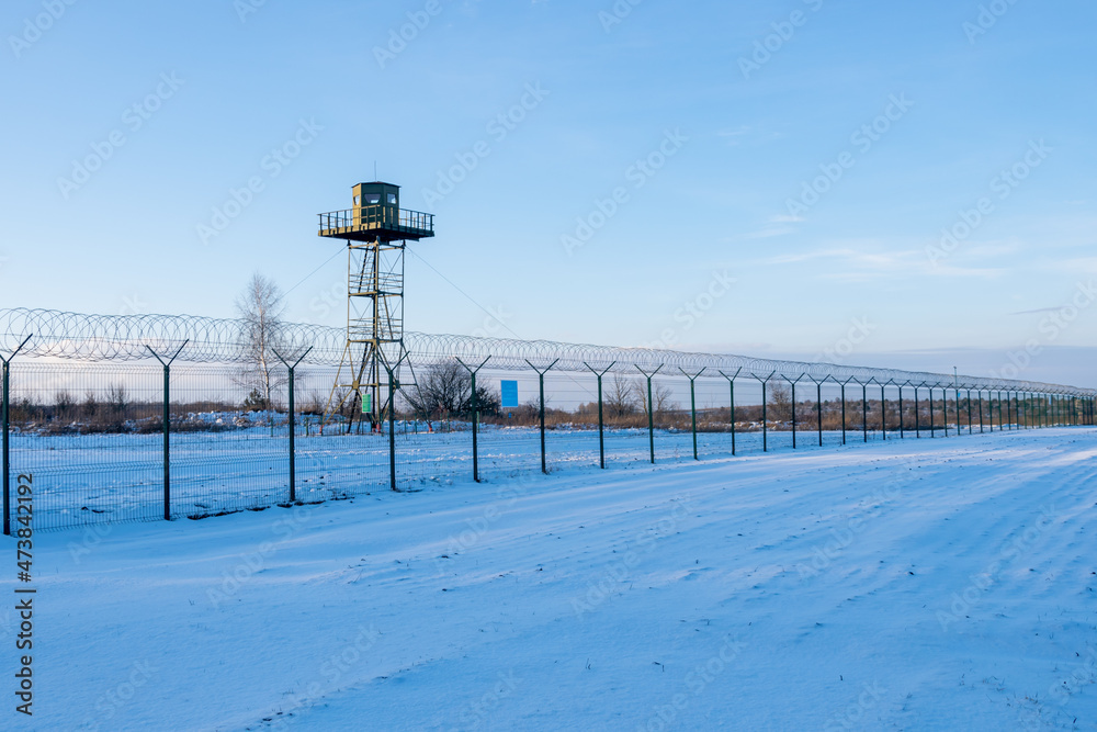 Border fence with barbed wire in front of the surveillance tower in a snow-covered field against a blue sky. Border protection and counteraction to illegal migration concept Border fence with barbed wire in front of the surveillance tower in a snow-covered field against a blue sky. Border protection and counteraction to illegal migration concept